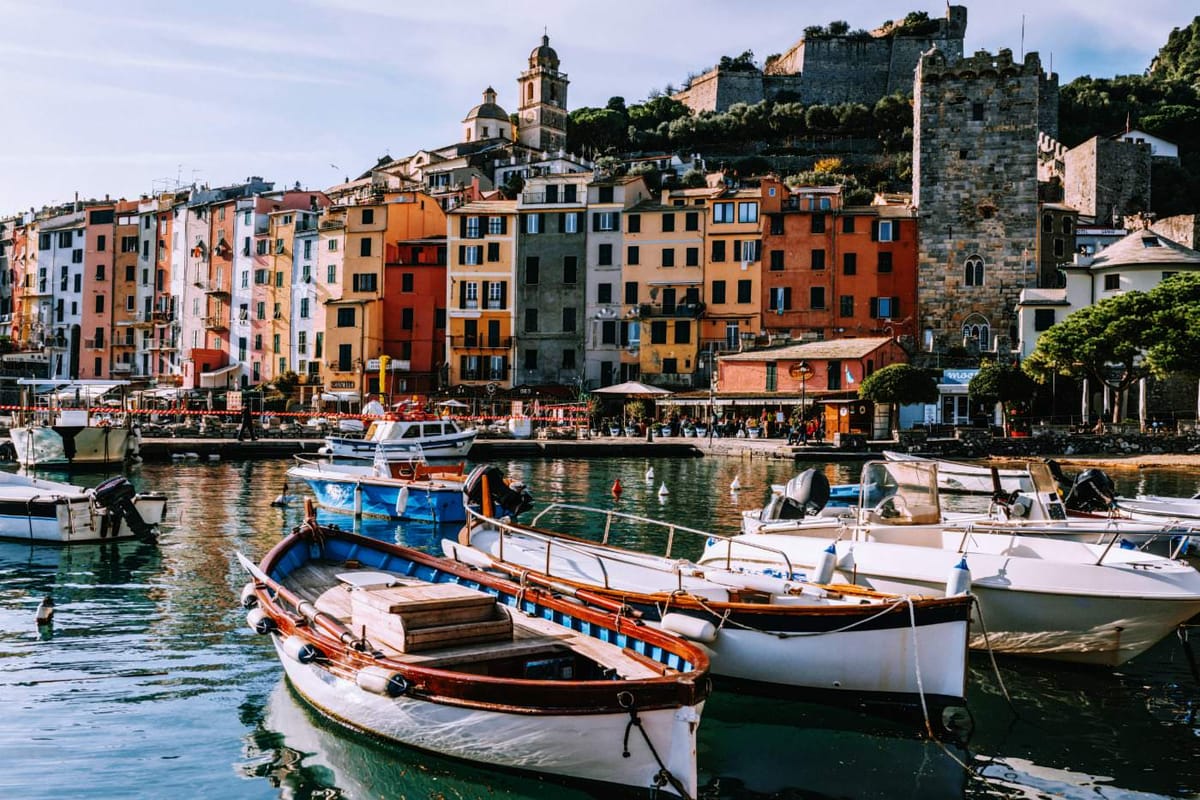 Colorful seaside town with boats in the harbor, vibrant buildings, stone tower, and a hillside church in Liguria, Italy.