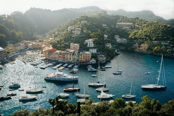 Aerial view of Portofino: colorful buildings and yachts in turquoise harbor, surrounded by lush green hills.